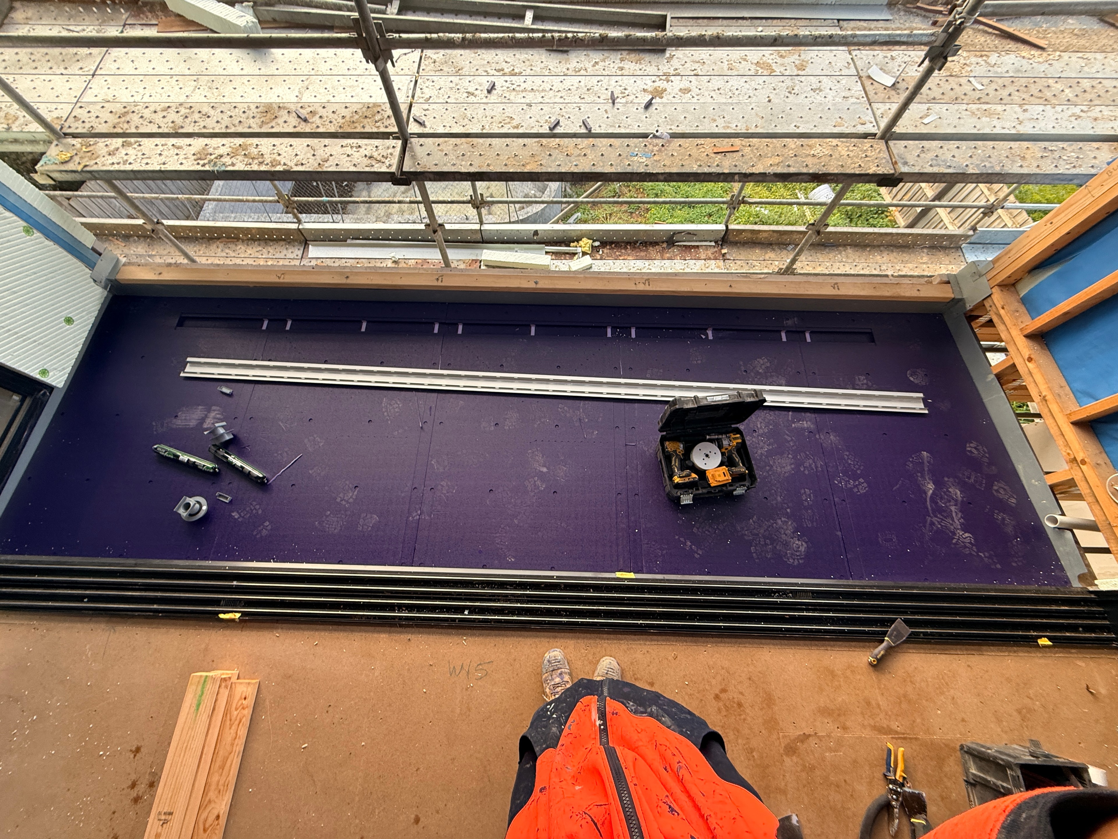 Balcony waterproofing in progress — purple membrane applied at threshold with adhesive bed being laid for tiling, scaffolding visible on new-build site in Dandenong by Casey Quality Coatings